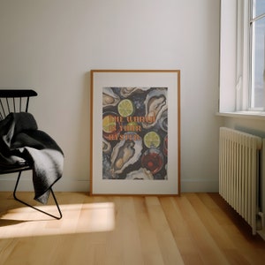 May include: A framed art print featuring an oyster and cocktail illustration. The artwork includes the text "The World Is Your Oyster" in orange and green. The print is displayed in a room with a chair and a window.