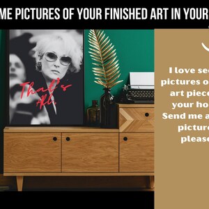 May include: A wooden dresser with two drawers and a black framed print of a woman with the text "That's All." The dresser is in front of a green wall with a gold leaf plant.