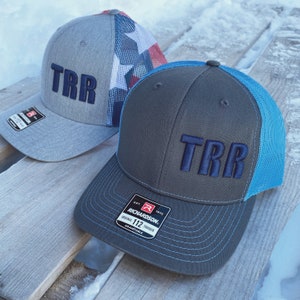 May include: Two gray and blue trucker hats with the letters "TRR" embroidered in navy blue on the front. The hat on the left has a red, white, and blue American flag design on the back. The hat on the right has a blue mesh back and a black label with the text "Richardson 112 Trucker Adjustable" on the side.