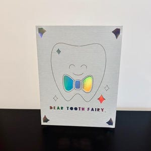 May include: A white card with a tooth illustration wearing a rainbow bow tie. The card says "Dear Tooth Fairy."