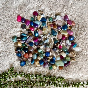 May include: A collection of polished gemstones in various colors, including pink, blue, green, and brown. The stones are scattered on a textured, light-colored surface, with a sprig of green foliage at the bottom.