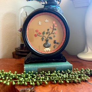 May include: A round, black framed artwork featuring a floral arrangement in a vase, with Chinese characters. The artwork is set on a green book and a wooden surface. A green, beaded plant is in the foreground.
