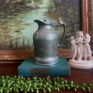 May include: A vintage pewter pitcher with a handle and lid, set on a teal book. The pitcher has a dark, aged appearance. A green beaded garland and a white sculpture of women are in the foreground, with a landscape painting in the background.