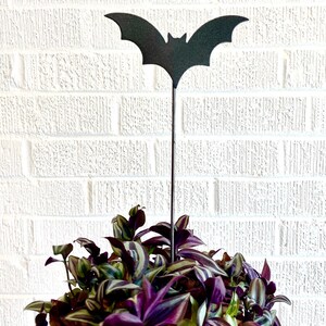 May include: A black metal bat garden stake with outstretched wings, placed in a pot of purple and green Wandering Jew plant. The bat stake is a decorative accent for Halloween or a gothic-themed garden.