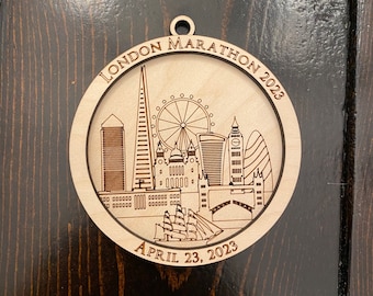 May include: A wooden ornament featuring a black and white illustration of the London skyline, including the London Eye, Big Ben, and the Shard. The ornament is inscribed with "LONDON MARATHON 2023" and "APRIL 23, 2023".