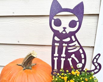 May include: A purple glittery cat skeleton cutout featuring large eye sockets and a detailed rib cage design. The cat figure is positioned next to a large orange pumpkin and a small potted plant with yellow flowers, creating a seasonal display.