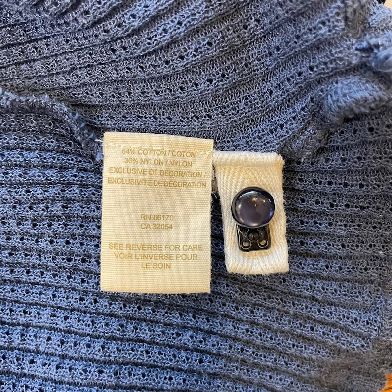May include: A beige fabric tag with black text that reads "64% Cotton/Coton 36% Nylon/Nylon Exclusive of Decoration/Exclusivite de Decoration RN 66170 CA 32054 See Reverse for Care Voir L'inverse Pour Le Soin".