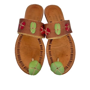 May include: A pair of brown leather slide sandals featuring a wide strap adorned with red and light green oval accents. Each sandal includes a matching light green toe post detail and a stitched border along the edge of the footbed.