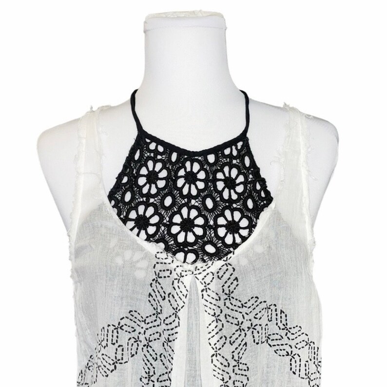 May include: White sleeveless top with black crochet detailing around the neckline. The top has a geometric pattern embroidered on the front.