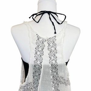 May include: White sleeveless top with a black embroidered design on the back. The top has a black tie at the neck.