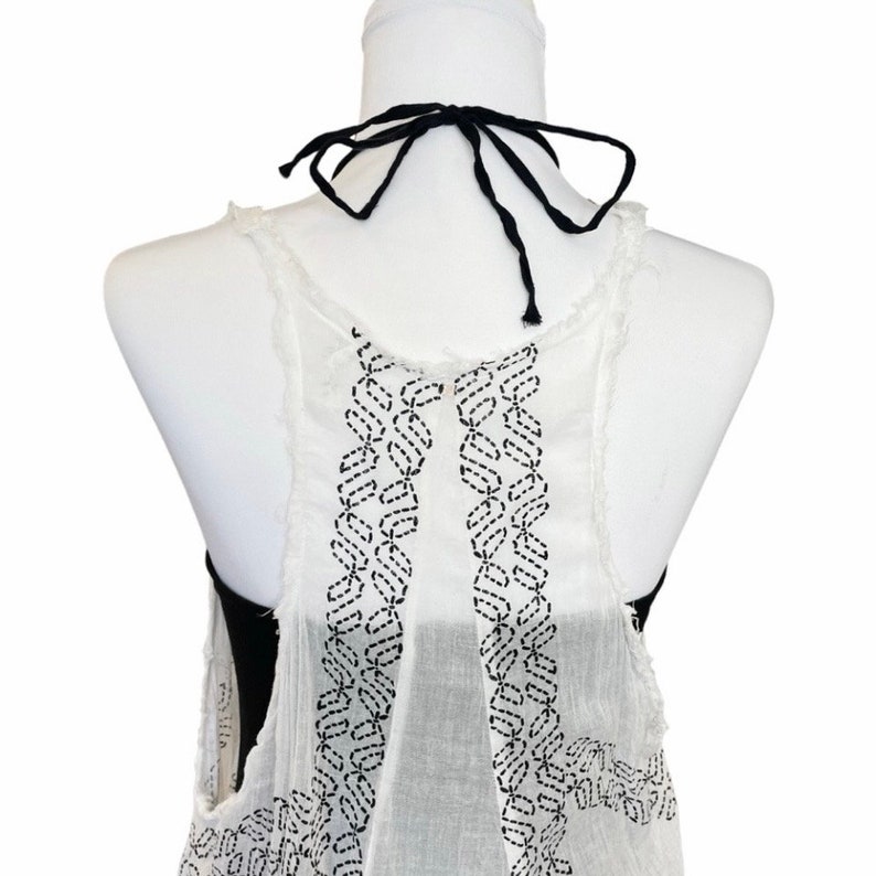 May include: White sleeveless top with a black embroidered design on the back. The top has a black tie at the neck.