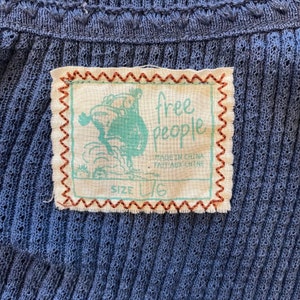 May include: A beige clothing tag with brown stitching around the edges. The tag features a cartoon image of a person riding a horse. The text "free people" is printed in black on the tag. The text "Made in China / Fait aux Chine" is printed in black below the logo. The text "Size L/G" is printed in black at the bottom of the tag.
