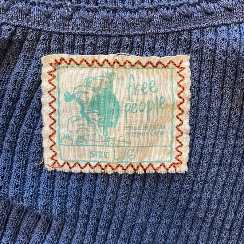 May include: A beige clothing tag with brown stitching around the edges. The tag features a cartoon image of a person riding a horse. The text "free people" is printed in black on the tag. The text "Made in China / Fait aux Chine" is printed in black below the logo. The text "Size L/G" is printed in black at the bottom of the tag.