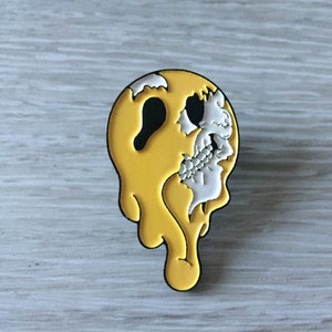 May include: A yellow and white enamel pin shaped like a skull with dripping yellow paint.