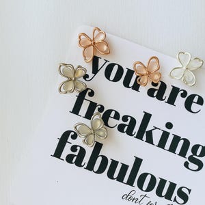May include: A collection of butterfly-shaped decorative pins in silver, rose gold, and white. The pins are arranged on a white card with the text "You are freaking fabulous".