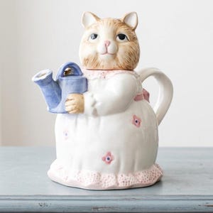 Könnte beinhalten: Eine keramische Teekanne in Form einer Katze, die eine blaue Gießkanne hält. Die Katze ist hellbraun und weiß und trägt ein weißes Kleid mit rosa Blumenakzenten und rosa Spitzenbesatz. Die Teekanne hat einen gebogenen Griff und einen Ausguss.