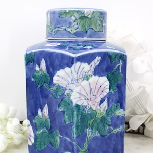 Könnte beinhalten: Eine blaue Keramikdose mit passendem Deckel. Die Dose ist mit weißen und rosa Blüten, grünen Blättern und Ranken verziert. Die Dose hat eine quadratische Form mit einem Deckel, der gut passt. Die Dose wird wahrscheinlich zur Aufbewahrung verwendet.