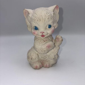 Peut inclure: Figurine de chat blanc vintage avec des yeux bleus, des joues roses et une bouche rose. Le chat est assis et lève sa patte. La figurine a une fourrure texturée et des accents roses sur les oreilles et les pattes.