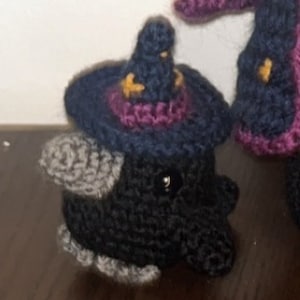 May include: Crocheted black creature wearing a dark blue witch hat with purple trim and gold stars. The handmade amigurumi figure has a gray ear and a button eye. A second purple and black figure is partially visible.