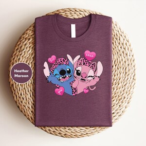 A purple t-shirt with a graphic design featuring two cartoon characters, a blue one and a pink one, along with hearts and the text "love you".