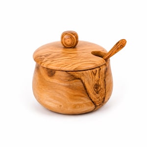 May include: A round wooden sugar bowl with a natural grain pattern, featuring a matching lid with a small knob handle and a cutout notch. A small wooden spoon rests inside the bowl, extending through the notch in the lid, showcasing the light brown and dark brown swirling wood texture.