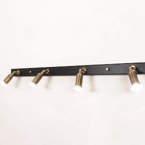 May include: A black track light with four brass-finished spotlights. The spotlights are adjustable and mounted on a black rectangular base. The fixture is attached to a white wall.