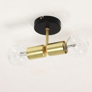 Può includere: Un moderno apparecchio di illuminazione a soffitto con base nera e accenti cilindrici color oro. Due lampadine trasparenti e rotonde sono attaccate all'apparecchio. La luce è montata su un soffitto bianco.