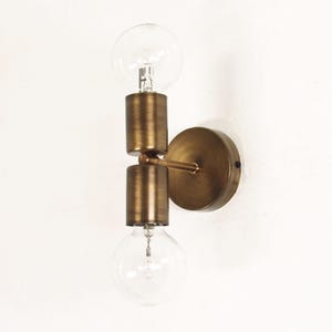 Può includere: Applique da parete in stile vintage con finitura in ottone. L'apparecchio presenta due lampadine trasparenti a forma di globo. Le lampadine sono posizionate sopra e sotto un corpo cilindrico centrale. L'applique è montata su una piastra rotonda.