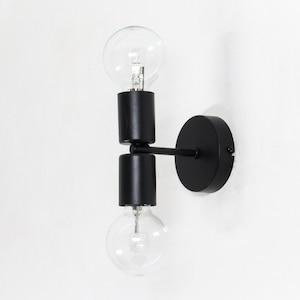 Può includere: Applique da parete nero moderno con due lampadine in vetro trasparente. L'apparecchio ha una base circolare e un braccio orizzontale. Le lampadine sono posizionate verticalmente, una sopra l'altra, e sono racchiuse in supporti cilindrici neri.
