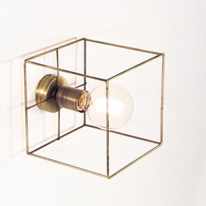 May include: A modern wall sconce featuring a clear, round light bulb encased in a geometric, cube-shaped frame. The frame and the base are made of a brass-colored metal. The light fixture is mounted on a white wall.
