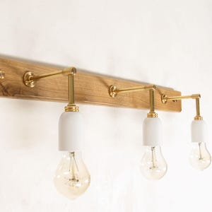 Peut inclure: Un luminaire en bois avec trois ampoules. Le luminaire a une finition en bois naturel et des bras en laiton tenant des abat-jour blancs et des ampoules de style Edison. Le luminaire est monté sur un mur blanc.