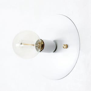 May include: A modern wall sconce featuring a white circular backplate, a gray bulb socket, and a clear, round Edison-style light bulb. A small brass accent is visible on the backplate. The fixture is designed for indoor lighting.