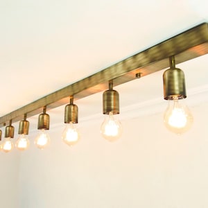 May include: A brass-toned, rectangular ceiling light fixture with six exposed light bulbs. The fixture is mounted on a white ceiling and has a vintage industrial design. The bulbs are illuminated, casting a warm glow.
