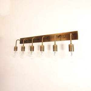 Peut inclure: Un luminaire de vanité à six lumières de couleur laiton. Le luminaire a une plaque arrière rectangulaire avec six bras incurvés tenant des ampoules en verre transparent. Le luminaire est monté sur un mur blanc.