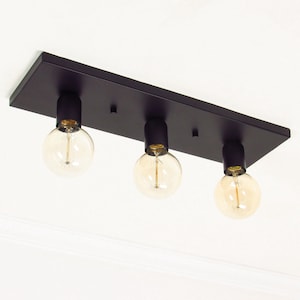 Pode incluir: Uma luminária de teto retangular preta com três lâmpadas redondas expostas. As lâmpadas têm um brilho amarelo quente. A luminária é montada em um teto branco.