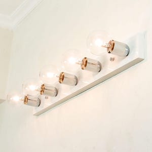 Peut inclure: Un luminaire de vanité rectangulaire blanc avec six ampoules claires et rondes. Chaque ampoule est enfermée dans une base métallique argentée avec un accent cuivré. Le luminaire est monté sur un mur blanc.