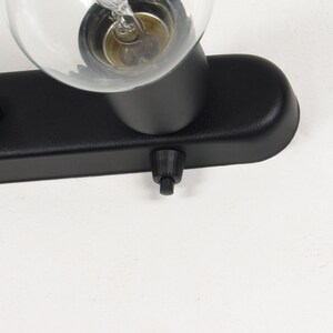 May include: Close-up of a black, rectangular light fixture with a clear glass light bulb. The fixture has a small switch and a cylindrical base. The light bulb is partially visible, showing the filament.