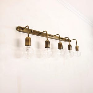Può includere: Un apparecchio per il trucco a cinque luci color ottone. Presenta una barra orizzontale con bracci curvi che sostengono lampadine rotonde e trasparenti. L'apparecchio è montato su una parete bianca, offrendo un'estetica vintage.