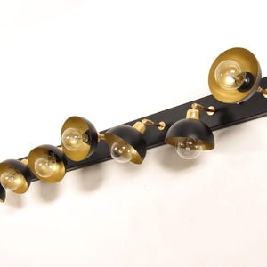 May include: A contemporary light fitting featuring seven lamps with black, dome-shaped shades and gold interiors. The lamps are mounted on a black rectangular base. The light bulbs emit a warm glow, creating a stylish and functional design.