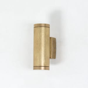 Pode incluir: Arandela de parede cilíndrica dourada com superfície texturizada. A arandela possui uma placa de montagem retangular e uma linha de destaque preta na parte superior e inferior. A luminária foi projetada para iluminação interna ou externa.