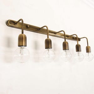 Op de afbeelding: Een messingkleurige vijf-lichte wastafellamp. De lamp heeft een rechthoekige balk met vijf gebogen armen, elk met een heldere, ronde gloeilamp. De lamp is aan een witte muur bevestigd.