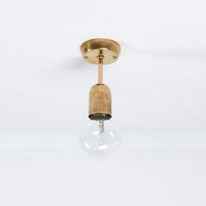 Pode incluir: Uma luminária de teto de latão com uma lâmpada de vidro transparente. A luminária tem uma base redonda e uma cúpula cilíndrica. A luz pende de uma haste. A luminária está em um fundo branco.