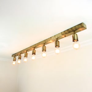 May include: A brass-toned, rectangular ceiling light fixture with eight exposed light bulbs. The fixture is mounted on a white ceiling, and the bulbs are lit, emitting a warm glow. The design is minimalist and industrial.