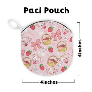 May include: A pink Paci Pouch with a repeating pattern of cartoon cows, strawberries, bows, and baskets. The pouch is 4 inches in diameter and has a white zipper and a white clip.
