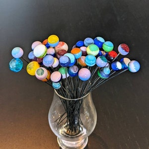May include: A clear glass vase holds a collection of colorful glass flower stems. Each stem features a round, marble-like head in shades of blue, yellow, pink, and white. The stems are black and the vase sits on a dark brown surface.