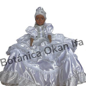 May include: A white doll wearing a white dress with lace trim and a white head covering. The doll has brown skin and black hair. The doll is sitting on a white background. The text "Botánica Okan Ifá" is visible in the background.
