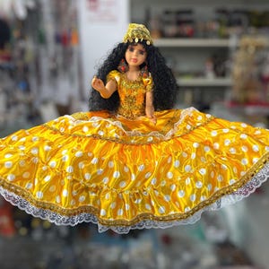 Op de afbeelding: Een pop in een gele jurk met witte stippen en gouden afwerking. De pop heeft zwart krullend haar en een gouden hoofdtooi. De jurk heeft een wijde rok en kanten rand. De pop is in traditionele stijl.