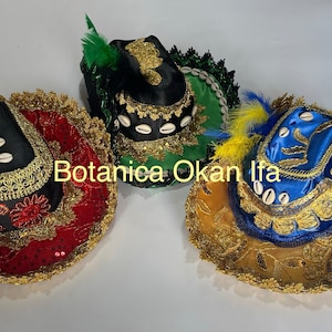 May include: Three traditional hats with gold trim and feathers. The hats are black, green, and blue with gold trim and white cowrie shells. The hats are on a white surface. Botanica Okan Ifa.