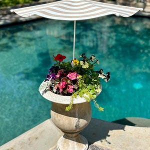 May include: A white and brown striped patio umbrella with a beige fabric canopy is positioned over a stone planter filled with colorful flowers. The planter is a light beige urn with a decorative base. The planter is positioned on the edge of a swimming pool.