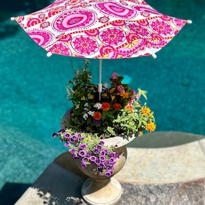 May include: A white patio umbrella with a pink and yellow paisley pattern, providing shade over a planter with purple, orange, and yellow flowers.